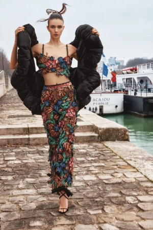 Fashion editorial shot of a woman wearing a floral two-piece outfit and black jacket, posing on a stone walkway near a river in Paris.