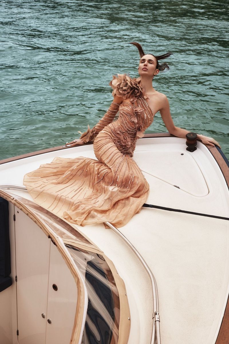 Woman in haute couture gown poses on a yacht in the ocean wearing stylized horns as a fashion statement; water background.