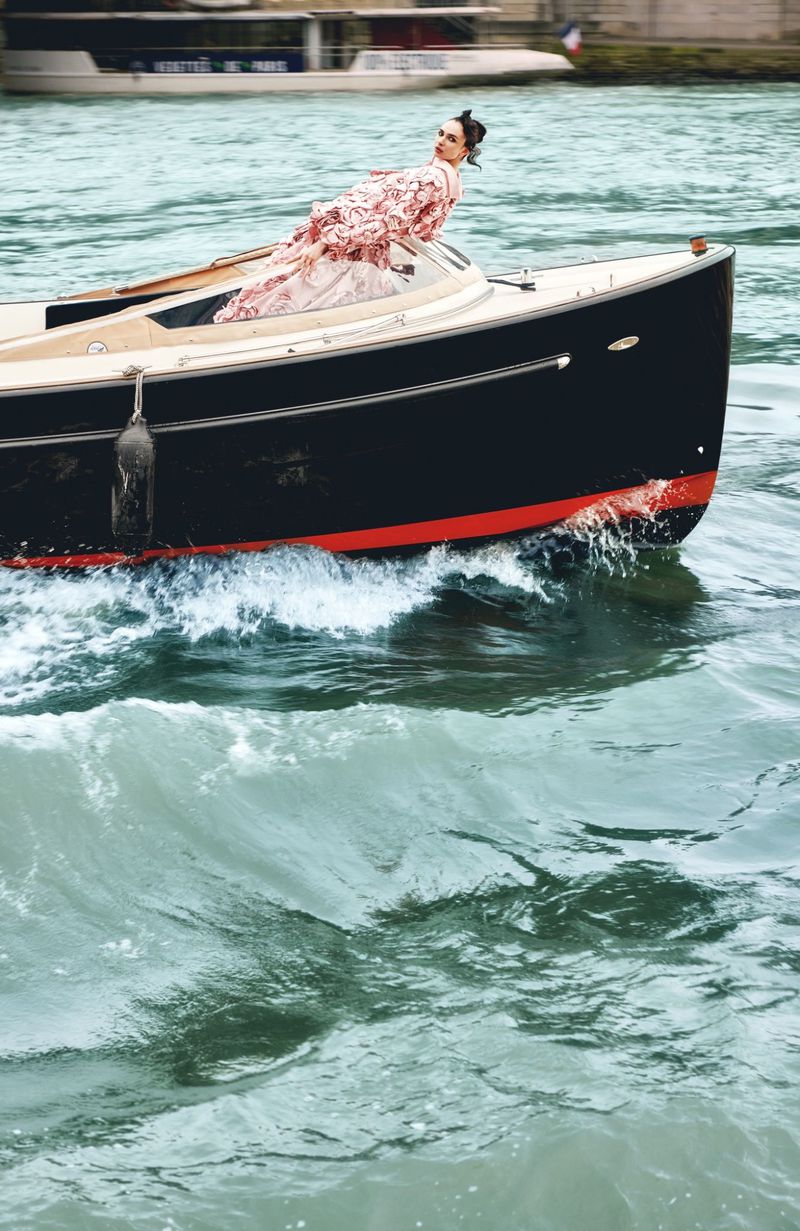 Model in a patterned gown leaning back aboard a dark motorboat creating waves on open water.