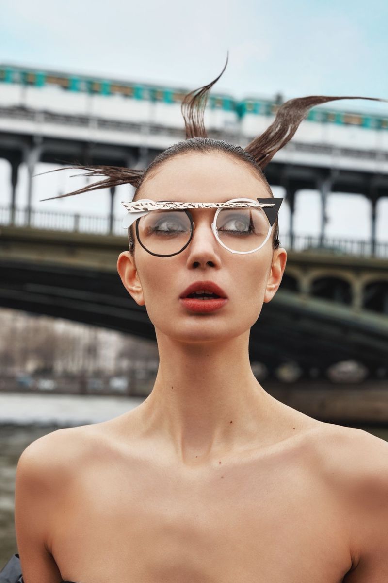 Adult female fashion model wearing decorative glasses posing in front of a city bridge with cloudy skies. Portrait photography.
