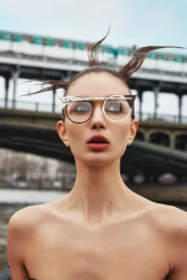 Model with avant-garde hair and sculptural eyeglasses against an urban bridge backdrop