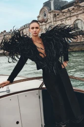 Model wearing a dramatic black feathered jacket posing on the deck of a boat with historic architecture and water in the background.
