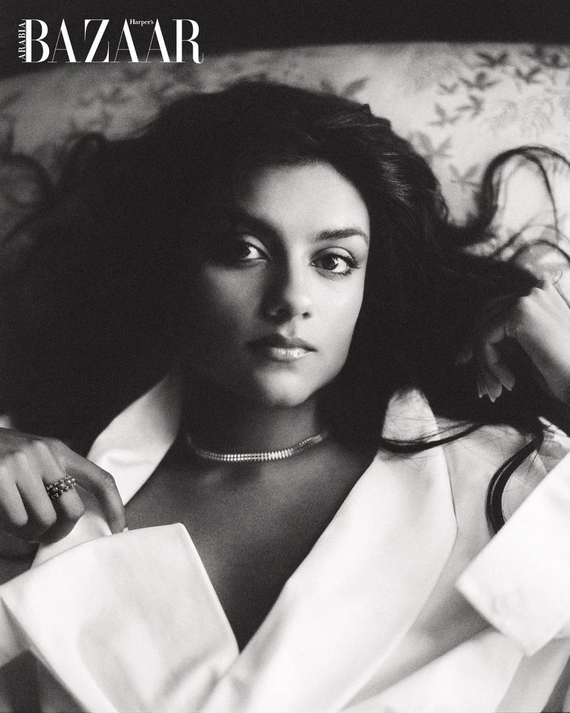Black-and-white portrait of a woman with dark wavy hair lying down wearing a white collared shirt, featured in Harper's Bazaar.