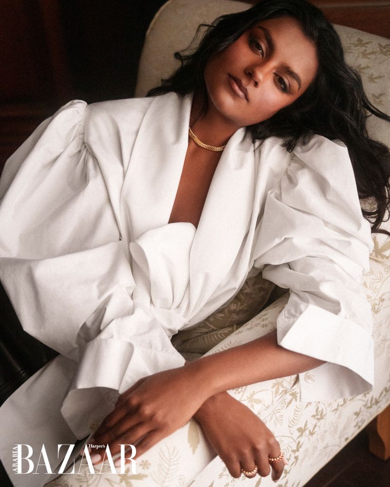 Simone Ashley in a voluminous white puff-sleeve top seated on an upholstered patterned chair for Harper's Bazaar Arabia