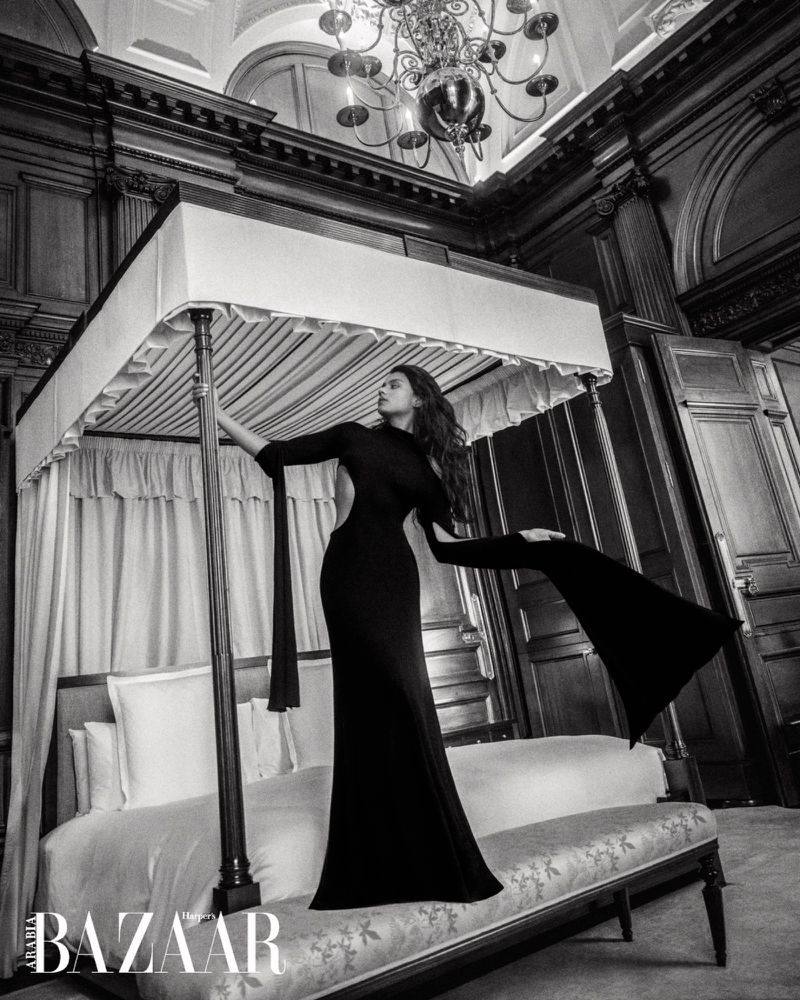 Simone Ashley in a black gown standing beside a four-poster bed in a luxury hotel room for Harper's Bazaar Arabia