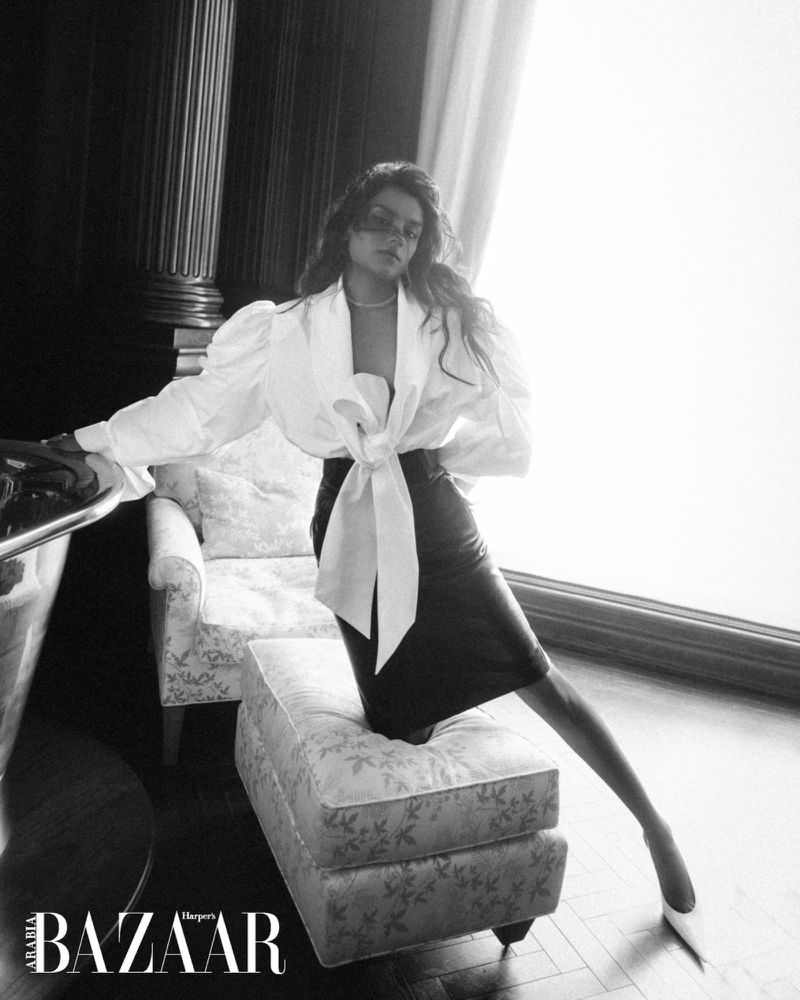 Monochrome fashion portrait of Simone Ashley kneeling on an upholstered ottoman in a white bow blouse and dark skirt
