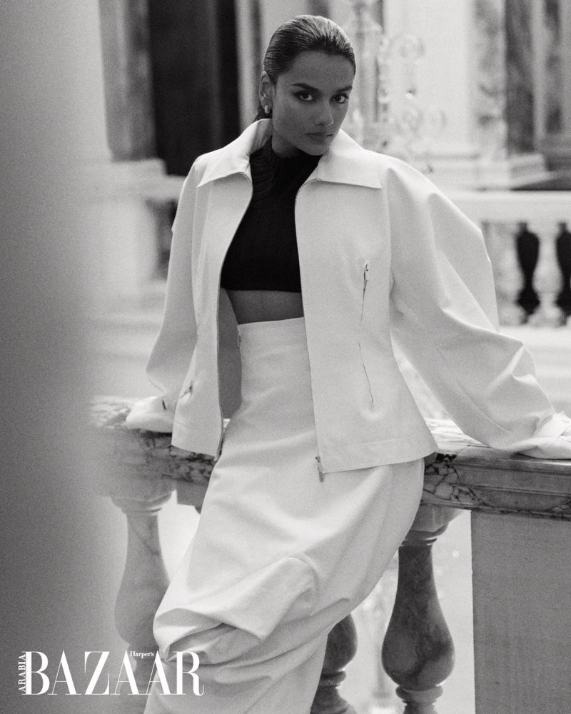 Simone Ashley leaning on a stone balustrade wearing a structured white jacket and skirt for Harper's Bazaar Arabia