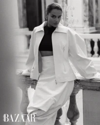 Simone Ashley leaning on a stone balustrade wearing a structured white jacket and skirt for Harper's Bazaar Arabia