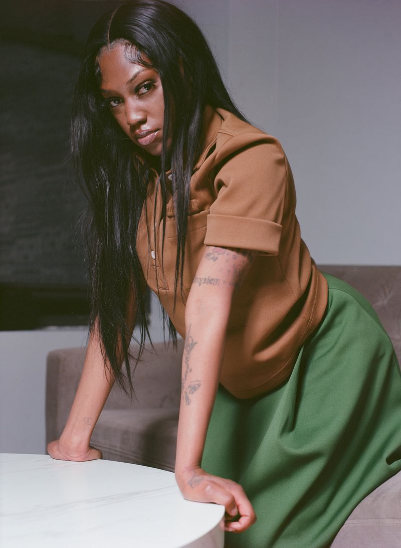 An adult female with long dark hair poses indoors, wearing a tan, short-sleeved polo shirt and a long, green skirt; casual fashion.