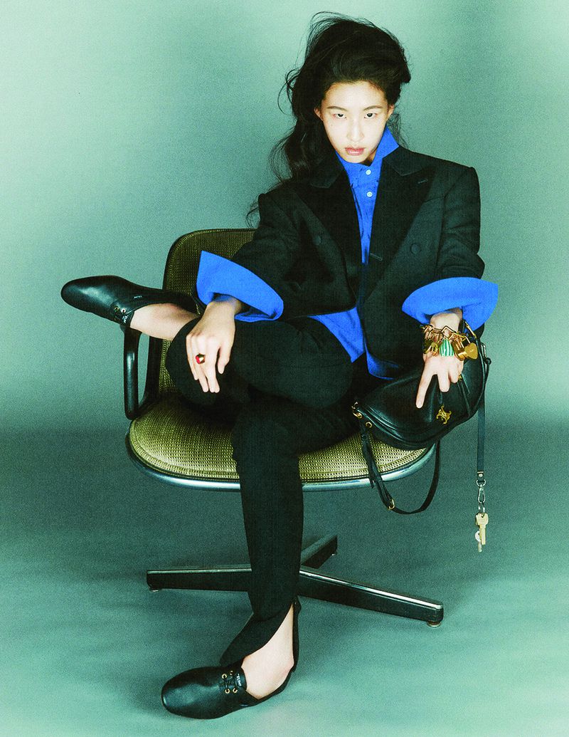 Model wearing a black suit and blue shirt seated on an office chair against a greenish backdrop for Elle Korea.