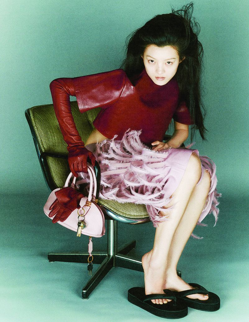 Asian woman wearing a burgundy leather top and feathered skirt, seated on an office chair holding a pink handbag