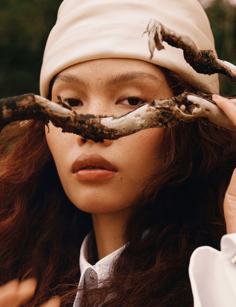 Close-up portrait of a model wearing a cream cap holding a gnarled tree branch in front of her face for Glass Magazine China ISLAND.