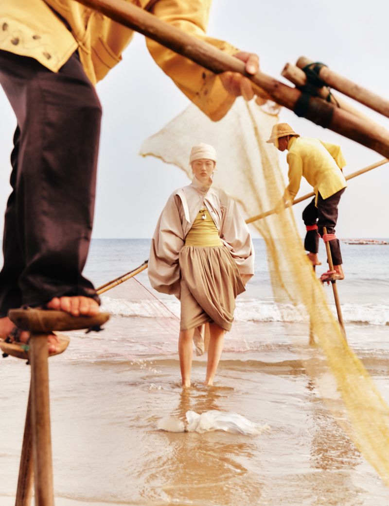 Model on a sandy beach wearing a beige draped outfit with oversized sleeves, framed by fishermen on stilts holding nets.