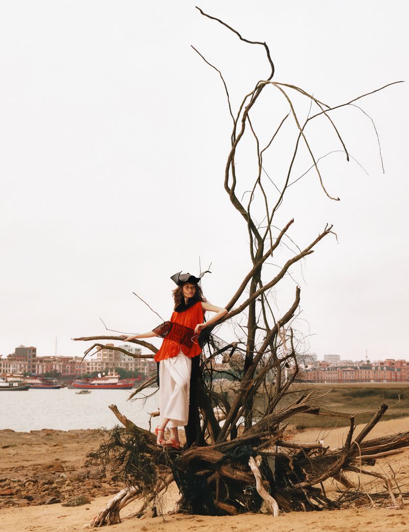 Model standing on a large dead tree trunk by a riverbank with a distant cityscape visible in the background