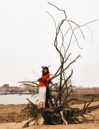 Model standing on a large dead tree trunk by a riverbank with a distant cityscape visible in the background