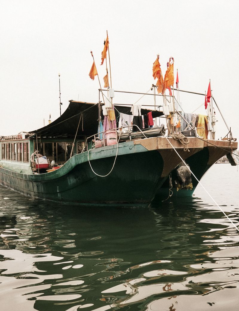 Traditional wooden green boat floating on dark water with orange flags and laundry hanging on deck