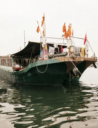Traditional wooden green boat floating on dark water with orange flags and laundry hanging on deck