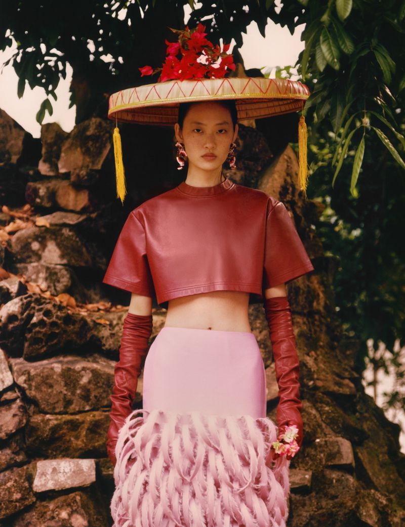 Model wearing a wide conical hat adorned with red flowers, a red crop top, and pink feathered skirt for Glass Magazine China ISLAND.
