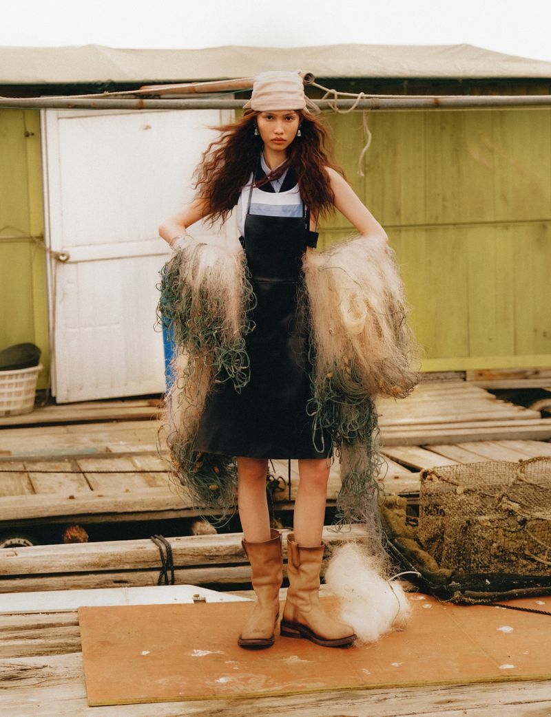 Model wearing a dark apron and tan boots holds large fishing nets standing on a wooden dock with a green building behind.