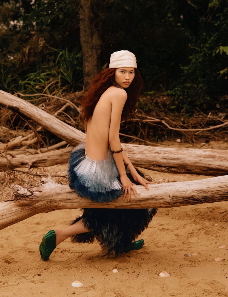 Model with long reddish-brown hair wearing a pale headwrap and feathered skirt posed on fallen logs on a sandy beach.