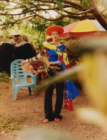 Model wearing a bright orange hat and colorful patterned top holding a basket of flowers outdoors near a blue plastic chair.