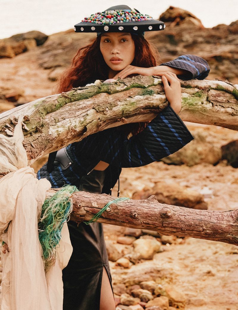 Model wearing an embellished flat hat leaning against a large driftwood log on a rocky coast for Glass Magazine China ISLAND editorial