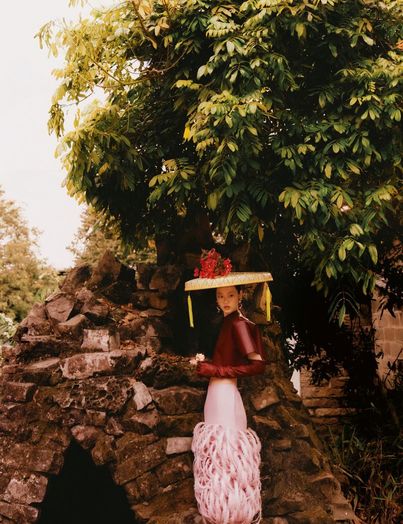 Model wearing a flat-brimmed hat, maroon top, and feathered pink skirt standing before a stone grotto beneath a tree