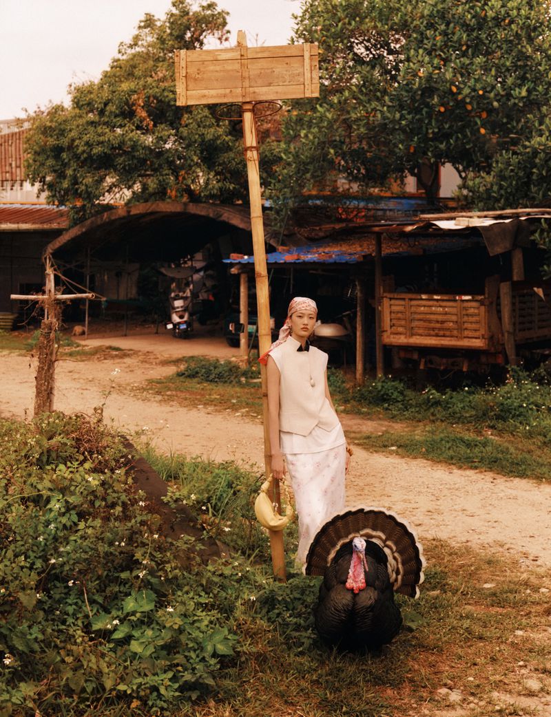 Model in a white vest and headscarf standing by a wooden pole with a turkey in the foreground outdoors.