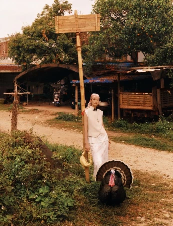 Model in a white vest and headscarf standing by a wooden pole with a turkey in the foreground outdoors.