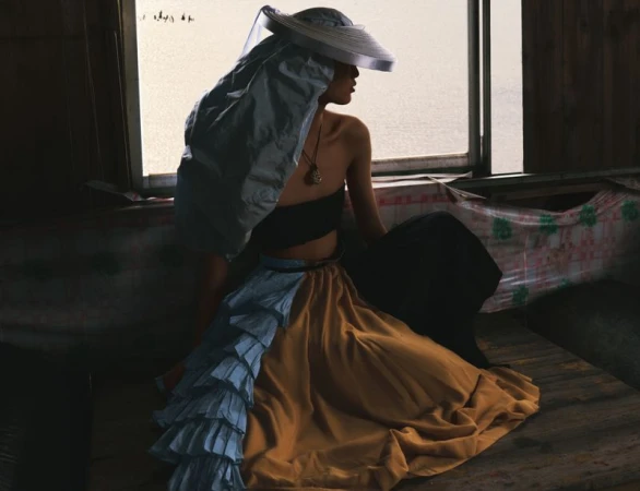 Model wearing a wide-brimmed hat with a draped fabric veil and a ruffled multi-tone skirt seated near a window for an editorial shoot