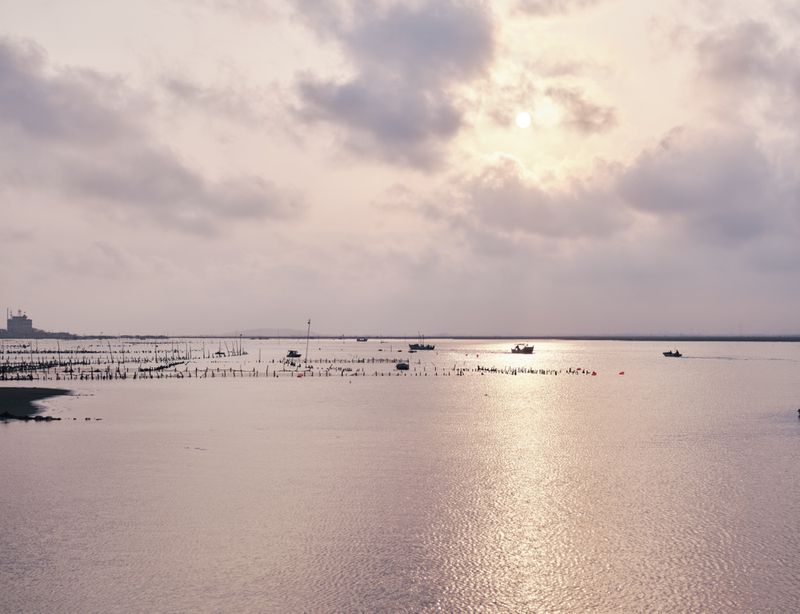 Wide tranquil coastal landscape with calm water, small fishing boats, and an overcast sunset sky with soft glowing clouds.