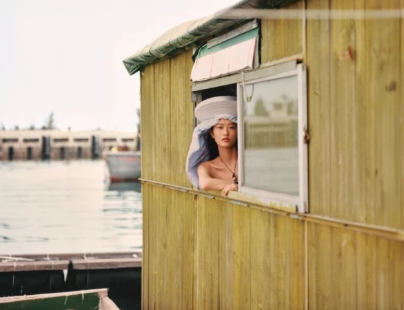 Model wearing a wide-brimmed hat looking out the window of a yellow wooden house on the water for Glass Magazine China.