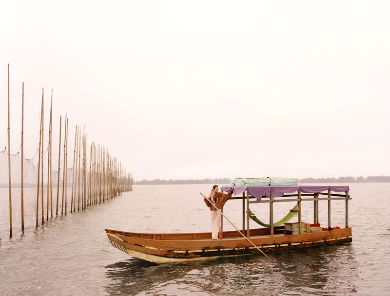 Model standing on a wooden boat holding a long pole on calm water next to a row of vertical wooden stakes.