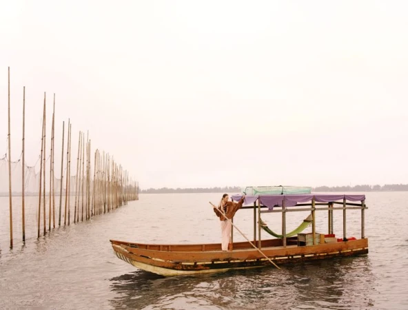 Model standing on a wooden boat holding a long pole on calm water next to a row of vertical wooden stakes.