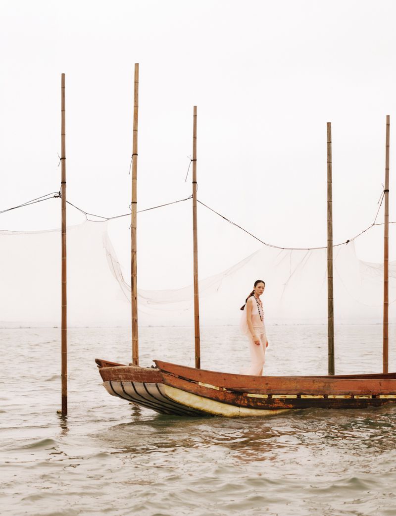 Model standing on a wooden fishing boat on the water with tall bamboo poles under an overcast sky.
