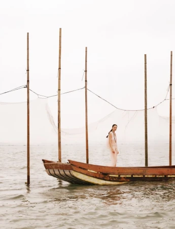 Model standing on a wooden fishing boat on the water with tall bamboo poles under an overcast sky.