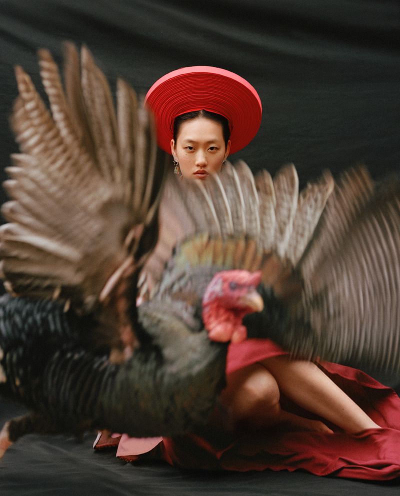 Model wearing a red hat and skirt seated behind a dark bird with blurred wings spread open in motion.