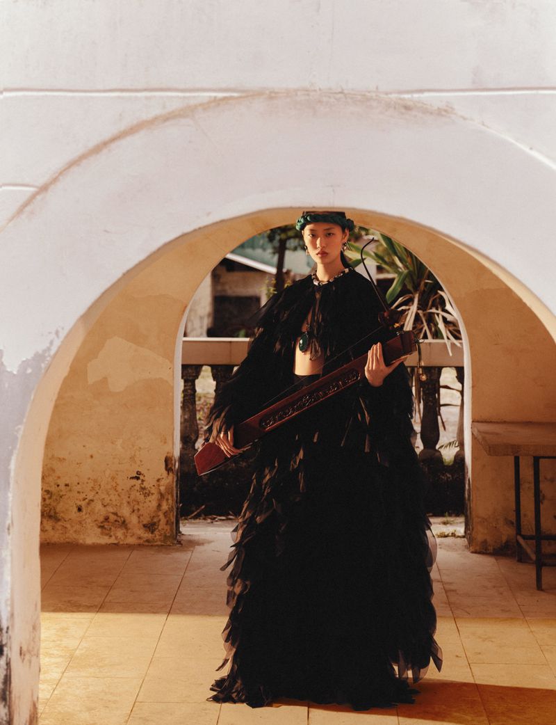 Model in a black feathered gown holding a long wooden instrument framed by a white arched colonnade