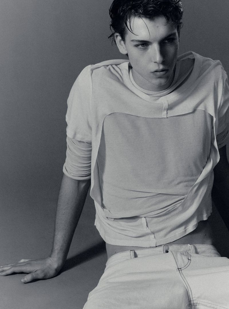 Young male model in a layered white t-shirt and light denim pants seated against a neutral grey background