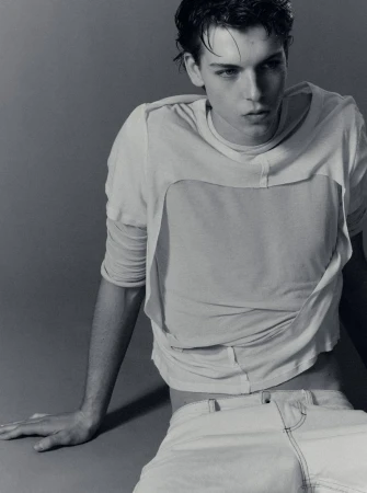 Young male model in a layered white t-shirt and light denim pants seated against a neutral grey background