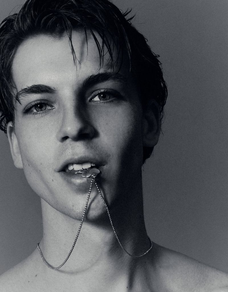 Close-up portrait of a young man biting a silver necklace chain against a neutral studio background