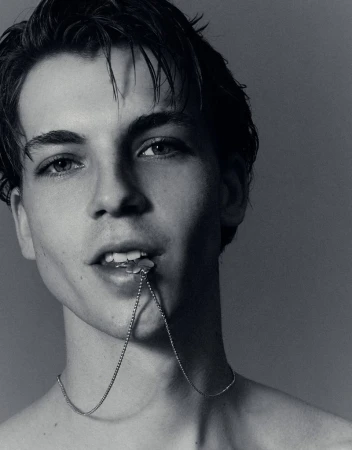 Close-up portrait of a young man biting a silver necklace chain against a neutral studio background