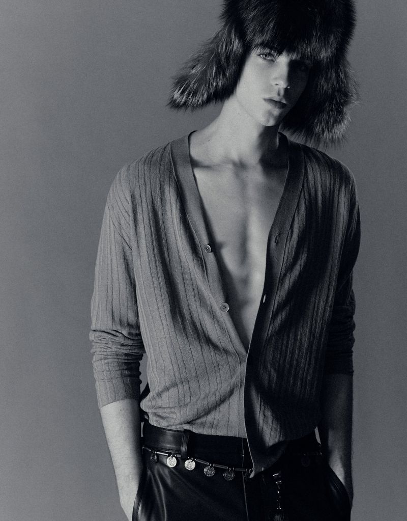 Monochrome portrait of a slim, young adult male with open cardigan, leather pants and furry hat, posing against a grey backdrop.