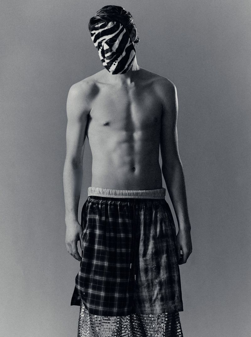 Shirtless male model wearing a black and white zebra-striped mask and plaid shorts against a neutral studio backdrop