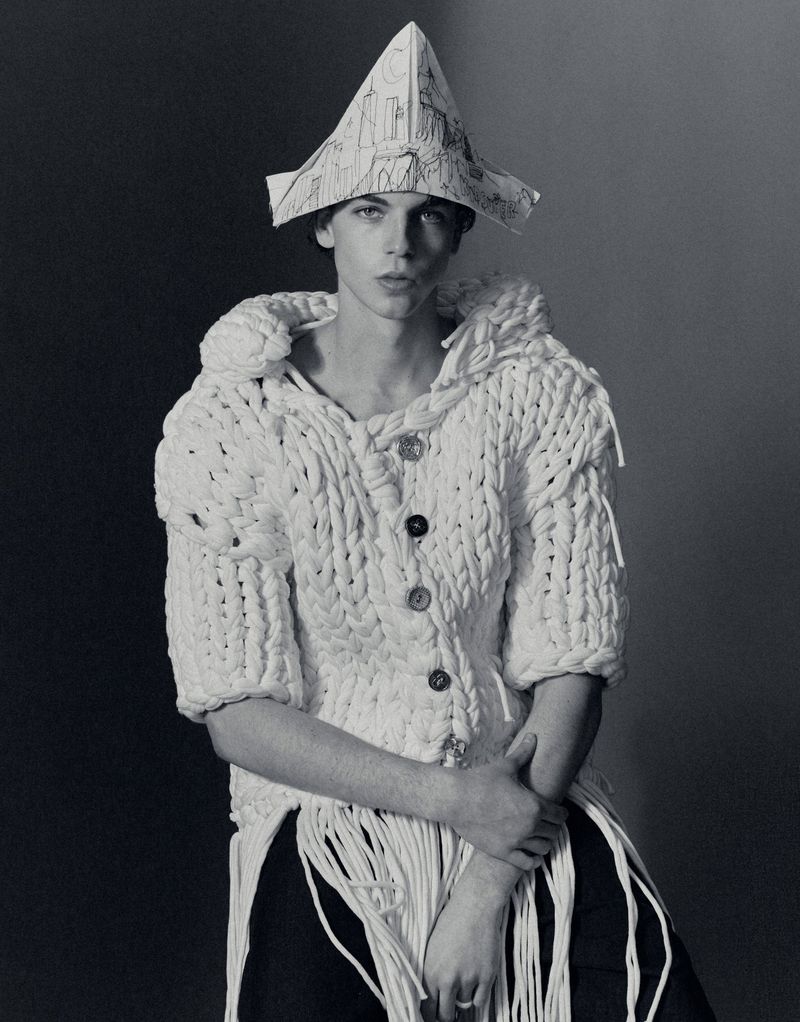Monochrome portrait of a male model wearing a chunky knitted white cardigan and a folded paper hat with sketches.