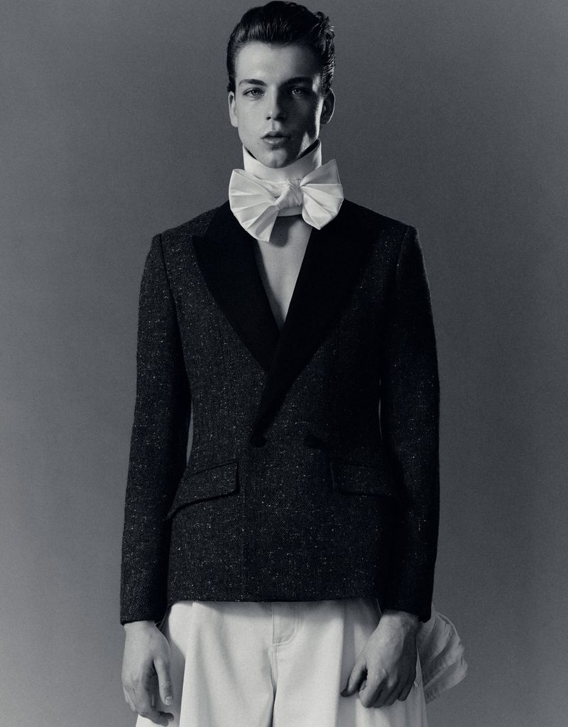 Model wearing a black tailored tuxedo jacket with a large white bow tie against a solid grey background