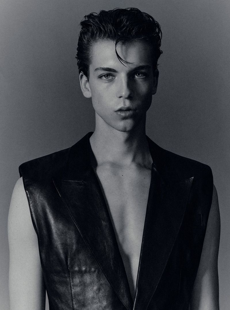 Portrait of a young male model with styled dark hair wearing a black vest against a plain grey studio backdrop.