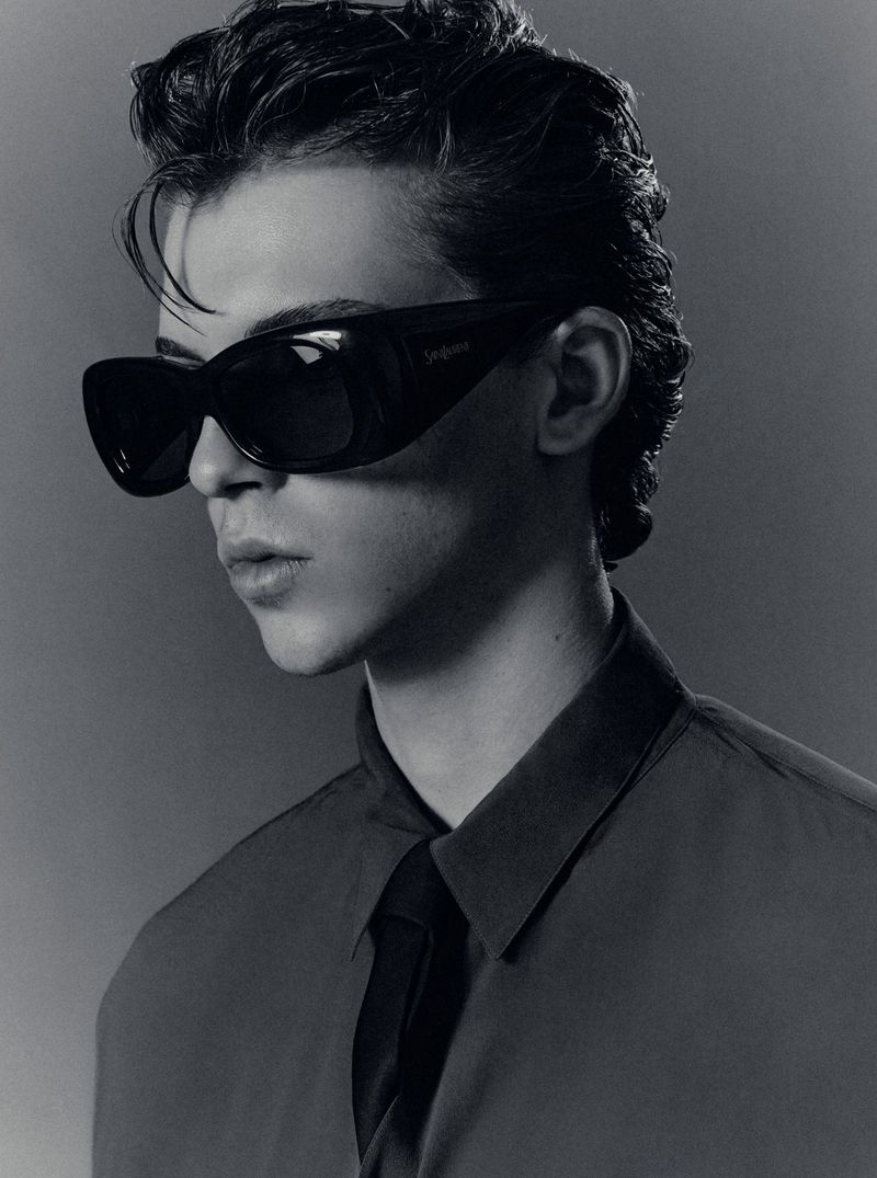 Portrait of a young man wearing dark sunglasses, a collared shirt, and a tie against a dark studio background