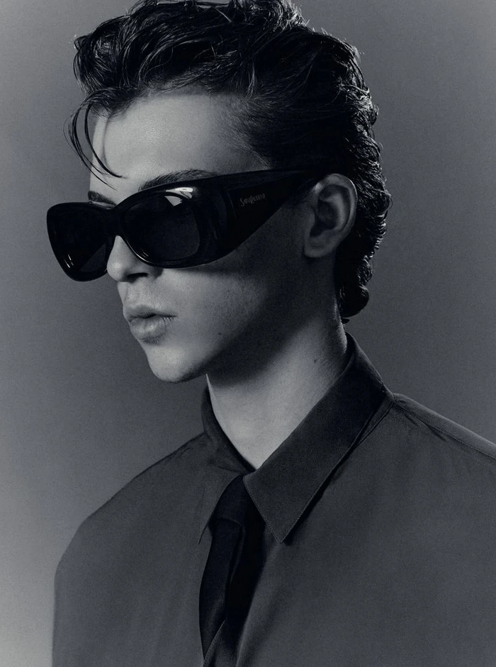 Portrait of a young man wearing dark sunglasses, a collared shirt, and a tie against a dark studio background