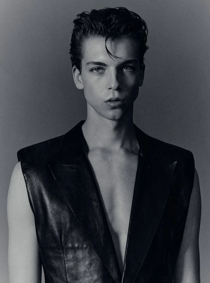 Portrait of a young male model with styled dark hair wearing a black vest against a plain grey studio backdrop.